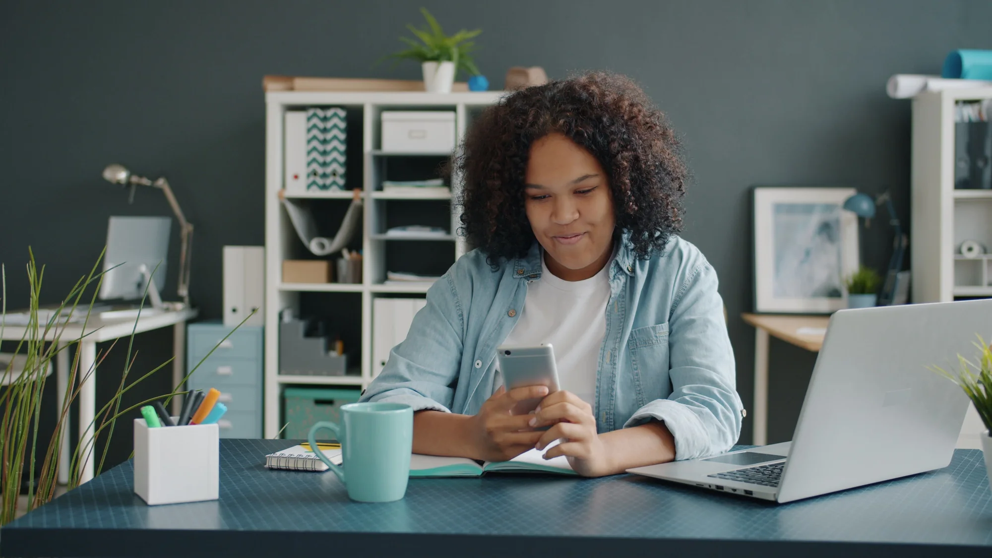 Eine Frau nutzt ihr Smartphone am Schreibtisch neben einem geöffneten Laptop.
